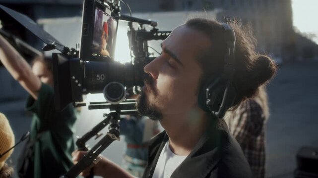 Close up view of focused cameraman in communication headset tracking scene on external field monitor displaying live take of actor on outdoor film set