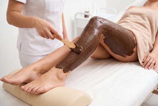 Spa therapist applying a luxurious chocolate mud mask to a woman's legs for a rejuvenating body treatment.