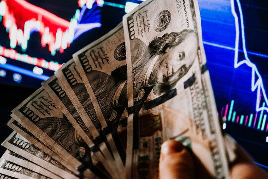 Hands holding US dollars currency in front of the stock exchange screen showing trading graphics.