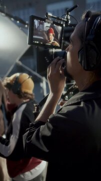 Male camera operator in headset watching field monitor screen while film director and actor discussing script on outdoor movie set. Vertical format video