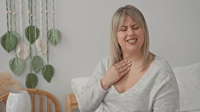 Woman coughing with hand to throat on bed wearing sweater and blonde hair, macrame wall visible in building; sickness recovery.