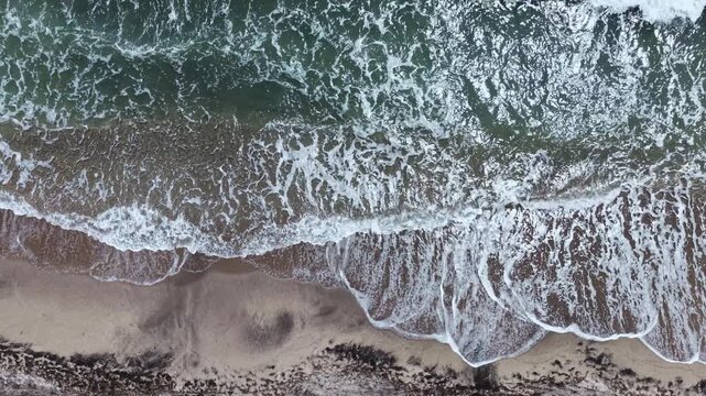 Waves roll onto the shore creating foamy patterns on the sand. The scene captures a part of the beach in late afternoon light. Water meets land in a continuous cycle.