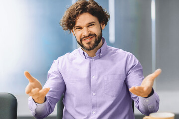 A man wearing a purple shirt talks and gestures with his hands in an office. He is seated at a...