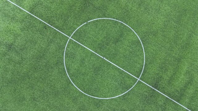 A soccer field shows the green grass with white lines marking the center circle and boundaries under clear sky in a local sports area. There are no players present.