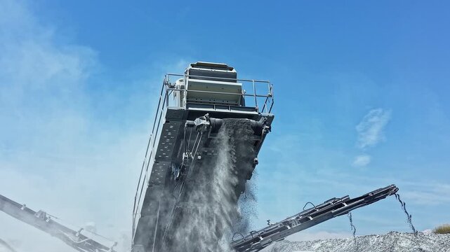 Large machinery in a granite quarry processes raw materials as dust rises into the air and blue sky provides a backdrop for daily operations.