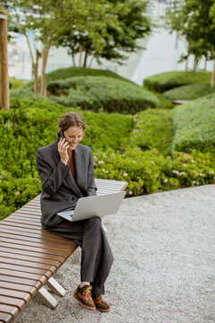 Woman works on laptop while talking on phone in a park setting during the day