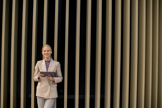 Woman stands with tablet outside a building with vertical lines during daylight hours
