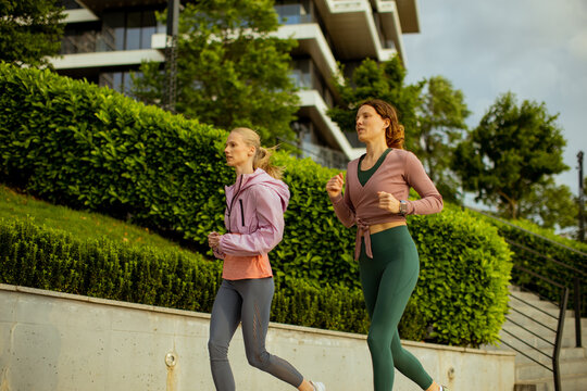 Women jog along a path in an urban area with buildings and green plants in view