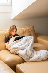 Obraz premium Blonde teenage girl drawing on tablet while resting under blanket on sofa at home.