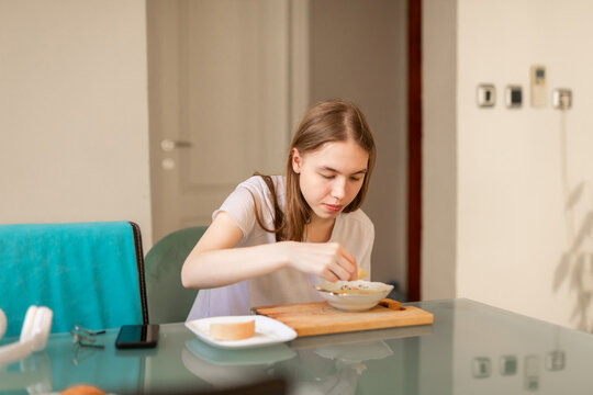 Blonde teenage girl in white shirt eats breakfast at glass table in bright home interior.