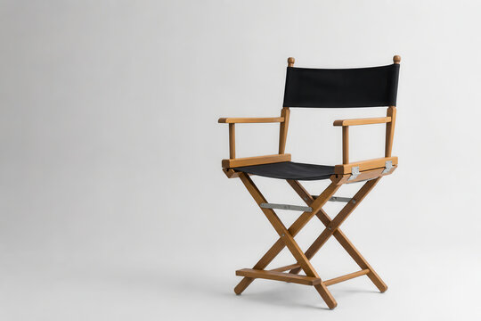 A wooden director's chair stands empty against a white backdrop. The chair has a black fabric seat and back. It is commonly used in film and photography settings.