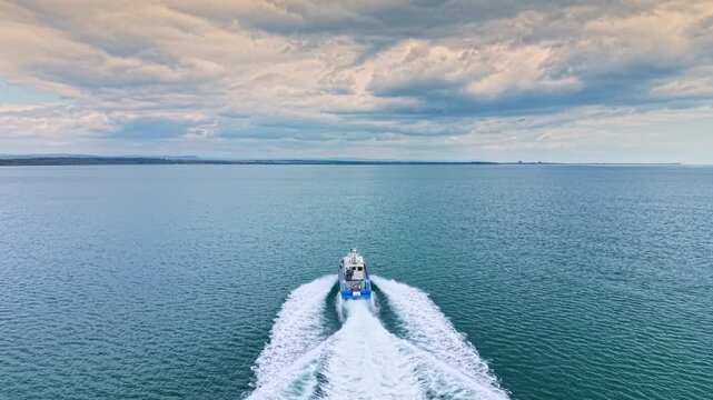 A fishing boat travels across the water in a coastal area. The scene shows waves and clouds above during late afternoon. The boat leaves a white trail behind as it moves.