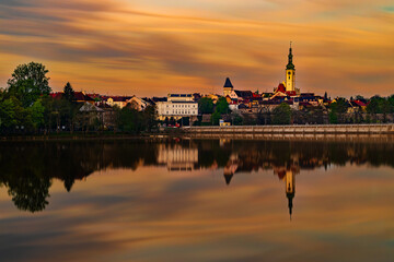 Obraz premium Historic town of Tabor in South Bohemia reflected in Jordan Reservoir at sunrise with Church of the Transfiguration on Mount Tabor beneath a colorful morning sky.