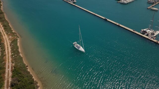 Aerial drone footage of a sailing yacht navigating crystal-clear waters near a marina and breakwater on Lefkada Island, Greece. Scenic Mediterranean coastline, turquoise sea