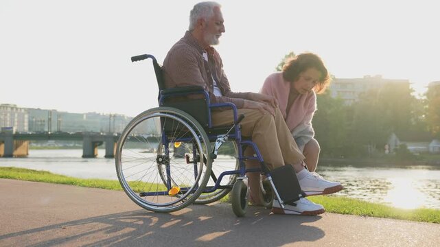 Old man in wheelchair walking with caregiver senior woman in park. Elderly family couple woman supporting taking care of paralyzed man in chair for people with disability outdoor. Rehabilitation