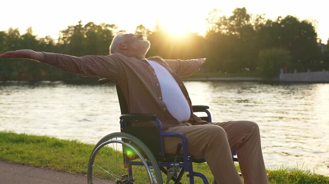 Happy senior man in wheelchair raised hands enjoying freedom walking on road in city park outdoor. Old happy man in chair for people with disability has a hope. Disability disease rehabilitation
