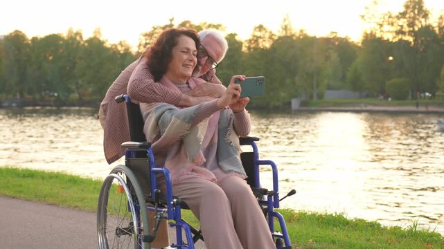 Senior couple woman in wheelchair caregiver old man making selfie enjoying walking. Elderly family man supporting embracing woman in chair for people with disability taking photo. Rehabilitation