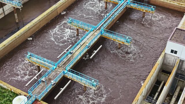 Close aerial aeration basin with machinery and bubbling surface, blue cross arms churn brown wastewater, metal walkway with visible pipes and valves, inspection