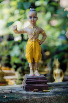 A statue of a young boy in traditional Thai clothing stands in Wat Umong temple in Chiang Mai, Thailand. The statue is adorned with gold jewelry and a yellow sarong.