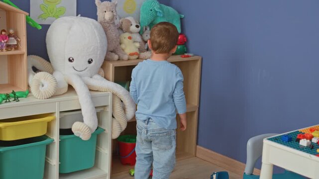 Boy picking up a yellow bucket next to a large plush octopus on shelves in a building playroom; curiosity play.