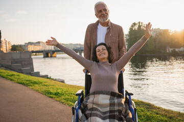 Fototapeta premium Senior woman in wheelchair walking with caregiver old man on road. Family couple man husband supporting taking care of paralyzed woman wife in chair for people with disability outdoor. Rehabilitation