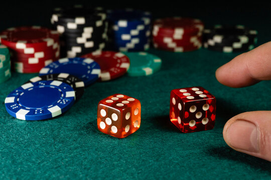 Two dice are rolled on a green felt table along with various colorful poker chips in the background. A hand reaches out toward the dice during a game