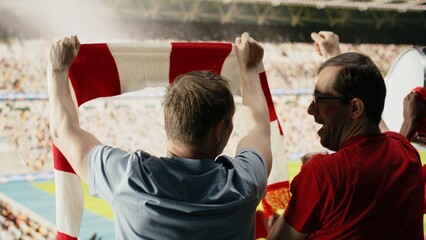Wide rear view of soccer fans in red shirts watching live football match from stadium stands with pitch and cheering crowd ahead and team spirit. © DC Studio