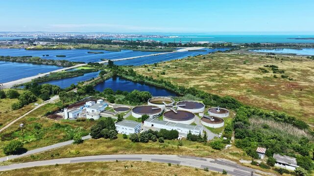 A water treatment facility is seen from above, situated near the coast. Surrounding land includes fields, rivers, and rows of solar panels under a clear sky.