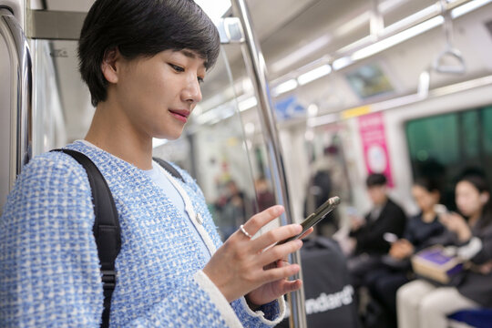 Young woman checks phone while commuting on subway. She wears stylish, textured jacket with backpack. Passengers sit quietly, focused on their own tasks. Soft lighting highlights her calm