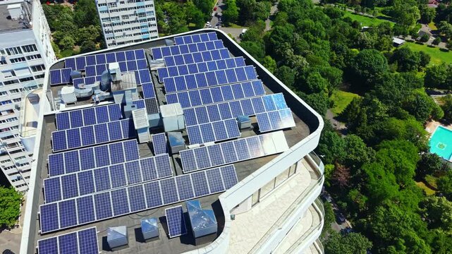 Solar panels are arranged neatly on a flat roof of a building. Surrounding the building are tall trees and green spaces. People may be nearby enjoying the outdoors.