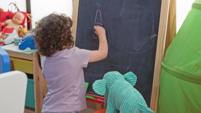 Girl drawing on a chalkboard, hand holding blue chalk and sketching a rocket beside a plush elephant and green play tent in a playroom; creative play joy.