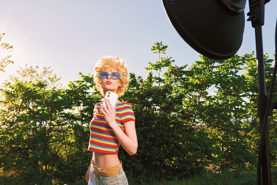 A young woman in a striped shirt and denim shorts stands outdoors, wearing blue sunglasses in sunlight, captured with a vintage color palette and carefree lifestyle vibe.
