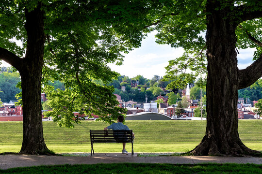 Man sitting on a park bench framed by mature shade trees overlooking historic downtown Galena, Illinois, on a summer evening