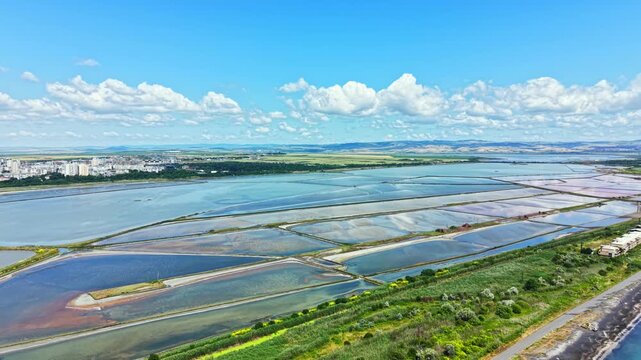 The scene shows salt pans in Burgas with varying water levels and colors. The sky is blue and filled with clouds. The city and hills can be seen in the background.