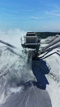 A granite quarry operation shows machines breaking down granite rocks. Dust is released into the air as conveyors move the rock powder. Surrounding hills are visible in the distance.