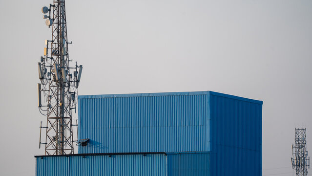 Telecommunication tower with antenna and dish beside blue industrial shed under grey sky