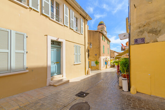 Sun-drenched ochre colored buildings with pastel shutters along a narrow cobblestone alleyway in the historic old town of Saint-Tropez, France, with the Chapelle de la Mis&eacute;ricorde in view.