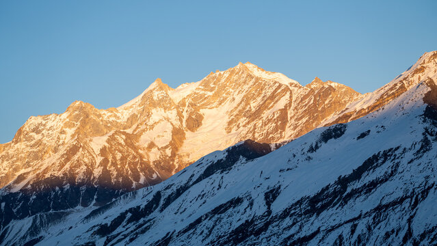 Golden sunrise light illuminating snow covered himalayan mountain range against clear blue sky