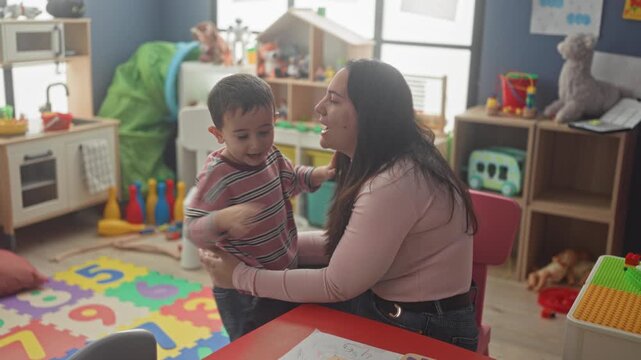 Woman seated hugging little boy on colorful foam number playmat in a daycare playroom inside building; affection bonding.
