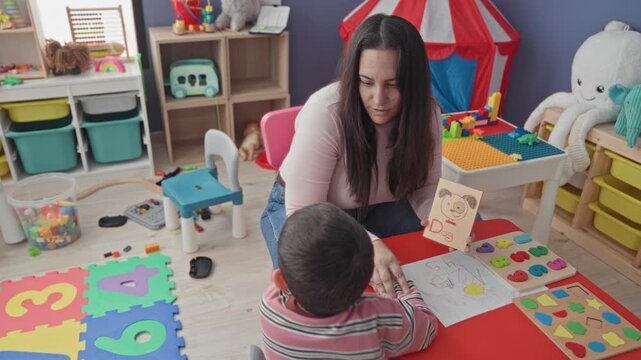 Woman holding a flashcard and offering a drawing while little boy points finger and reaches across a red table amid toys and foam numbers in a playroom building; concentration.