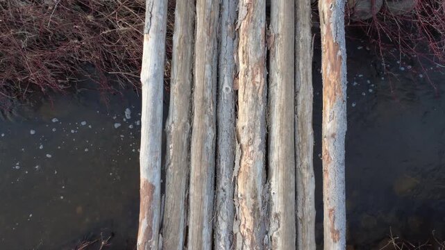 Drone footage rises above a rustic wooden log bridge crossing a bubbling stream near an old skidder and muddy road leading to yellowing agricultural fields.