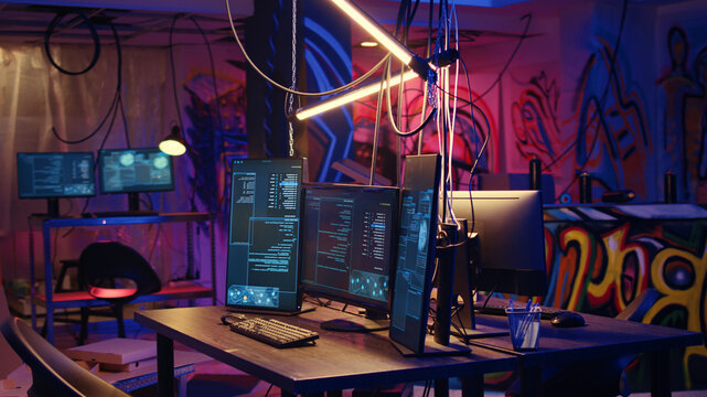 Panning shot of high tech computer system running malicious code in empty basement, PC monitors in empty neon lit criminal hideout used by hackers to commit illegal activities.
