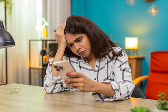 Indian woman at home views bad news on smartphone frowning presses button and sets device aside feeling very upset. Arabian girl at table shows disappointment negative emotion shaking head after loss.