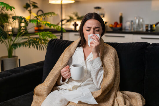 Young woman at night home under blanket with hot tea and medicine sips for sore throat cough. Caucasian girl on sofa at evening uses tissue sneezes feels fever fatigue so rests quiet asks help softly.