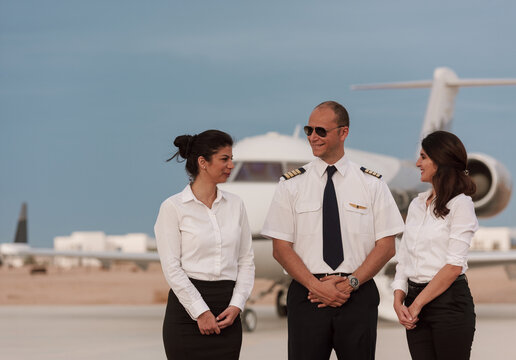 Friendly private jet crew with smiling pilot and two flight attendants in white uniforms talking on airport tarmac at golden dusk beside a business jet ideal for luxury aviation and charter travel