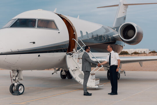 Airline pilot in uniform greeting a businessman with a friendly handshake beside private jet stairs on airport tarmac under soft overcast sky ideal for luxury business travel and charter service