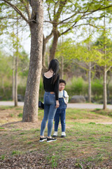 Fototapeta premium Mother and child stand beneath a tree. They gaze forward, sharing quiet moments. Soft sunlight filters through green leaves. Natural park setting with grass and paths. Ideal for family, parenting