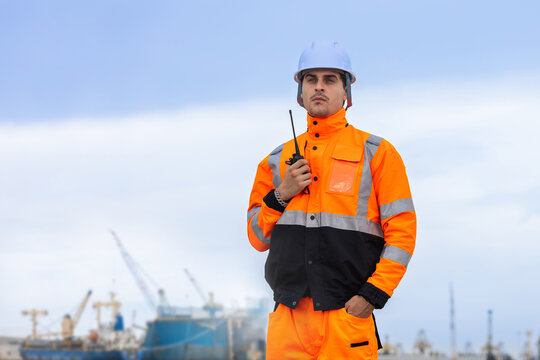 Portrait of a professional port supervisor using a walkie-talkie at a busy shipping terminal, Logistics coordinator in high visibility uniform overseeing harbor operations