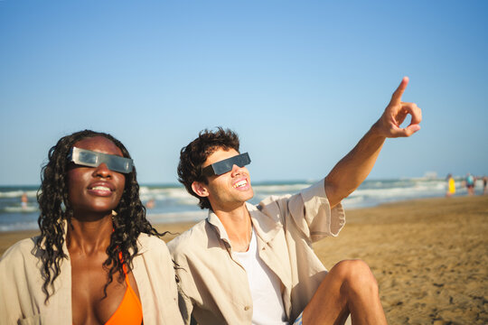 Two people on beach wearing solar eclipse glasses, man pointing to sky