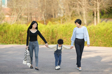 Obraz premium Three people stroll together on a paved path. Woman and child hold hands, smiling softly. Another woman walks beside them, relaxed and content. Trees stand bare in the background